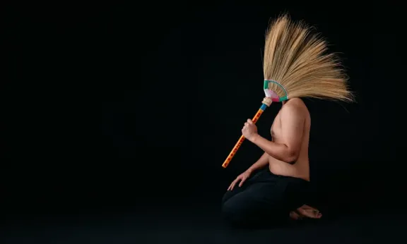 A person sitting on their knees against a black background holding a traditional Filipino broom that covers their head, acting as a headdress.
