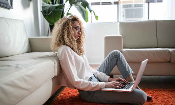 A student sitting next to a sofa and using a laptop