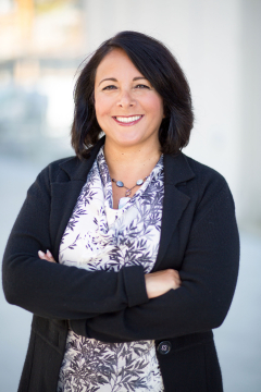 Dark-haired woman, smiling, standing with arms crossed in front of a light building. 