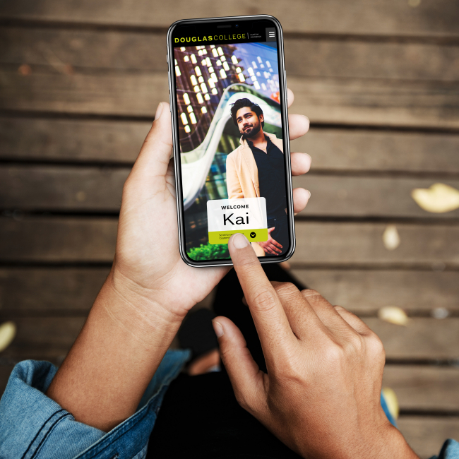 A person is holding a mobile phone with two hands. One finger is touching the screen of the phone. On the screen there is a photo of a young man and the words Welcome Kai.